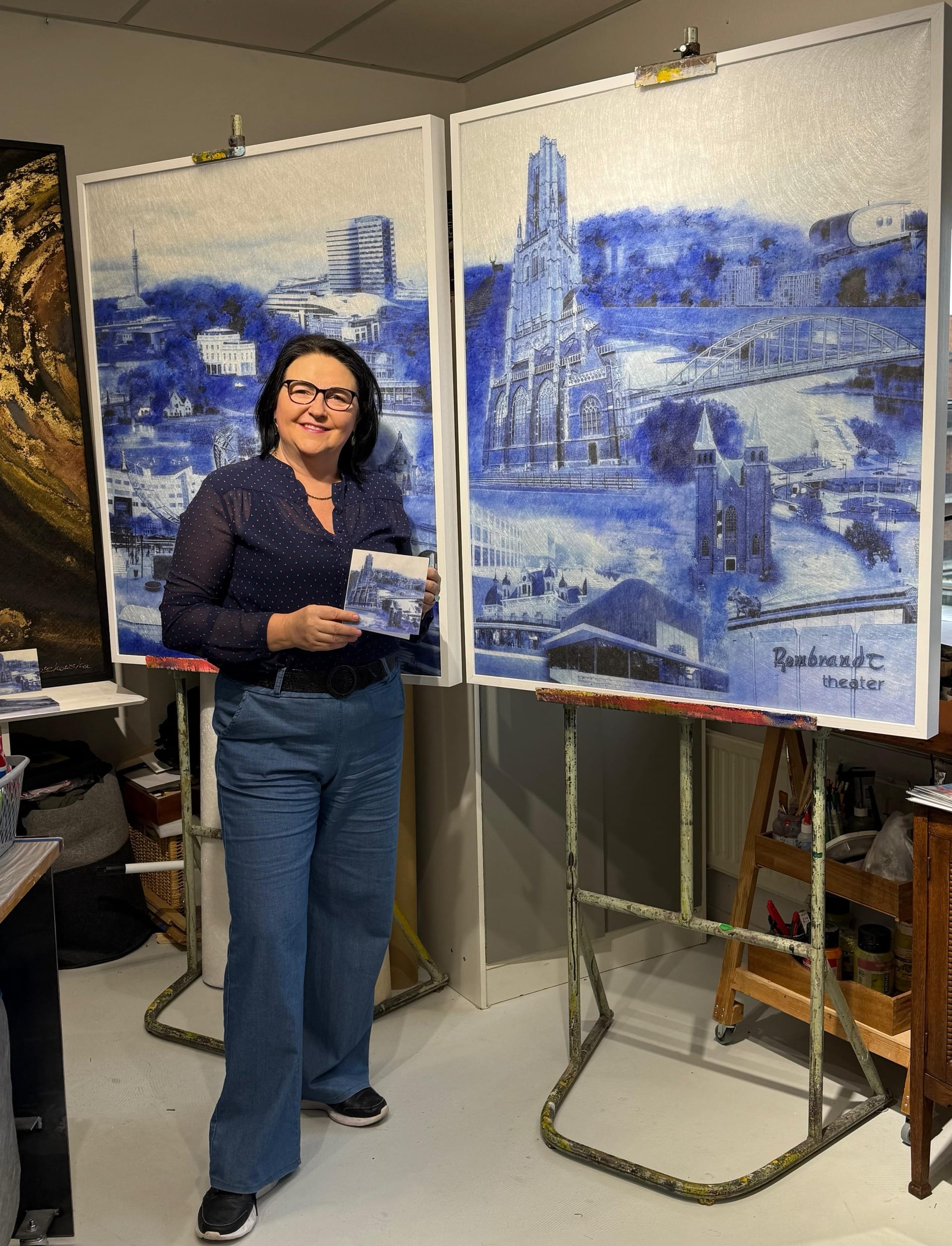 Dorota Roszkowska in haar atelier, tussen twee grote blauw-witte schilderijen van Arnhemse gebouwen, met een kaart van hetzelfde werk in haar handen.